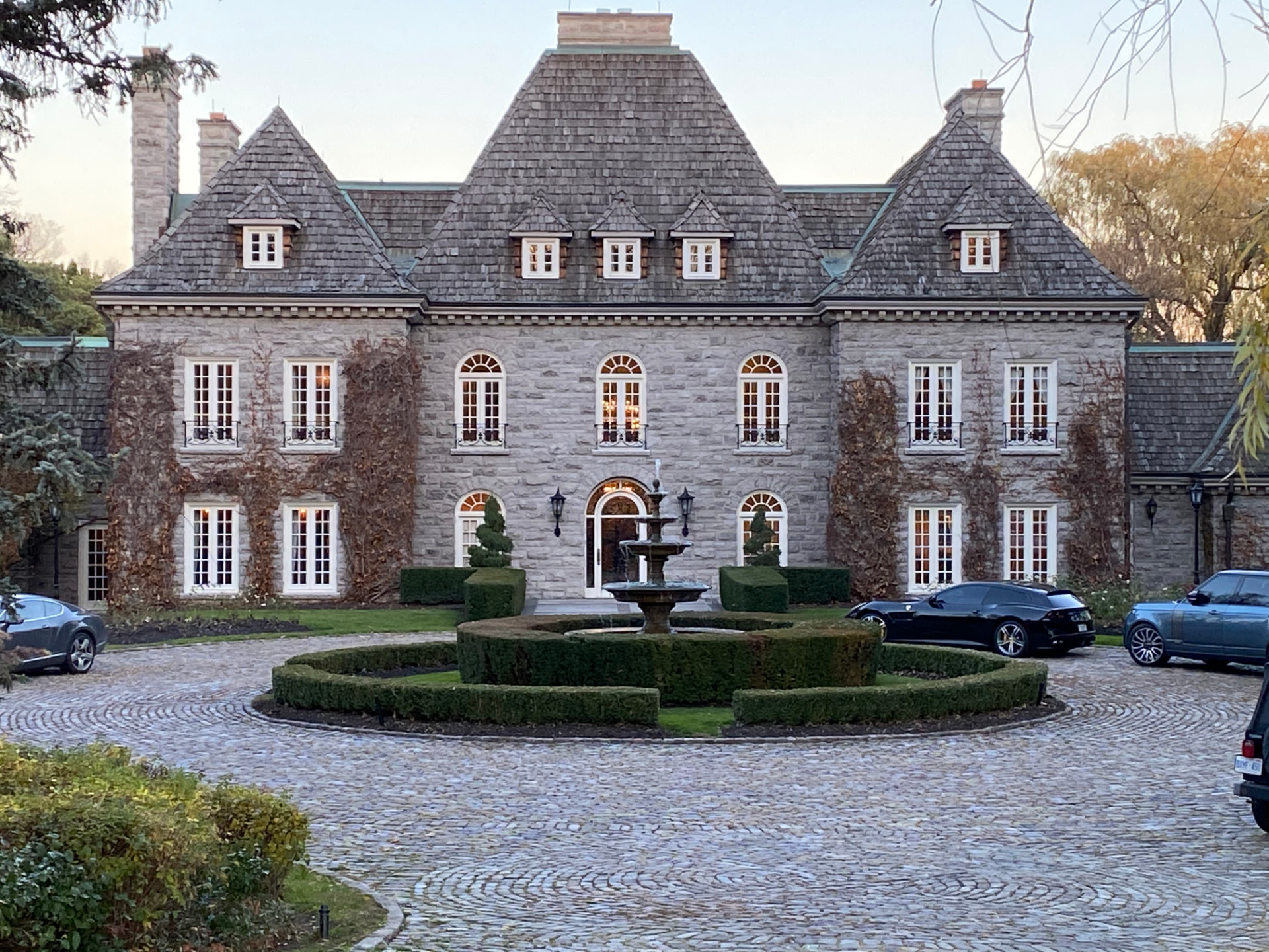 Symmetrical French Chateau-style stone mansion at dusk, with a tiered fountain at the centre of a circular cobblestone driveway